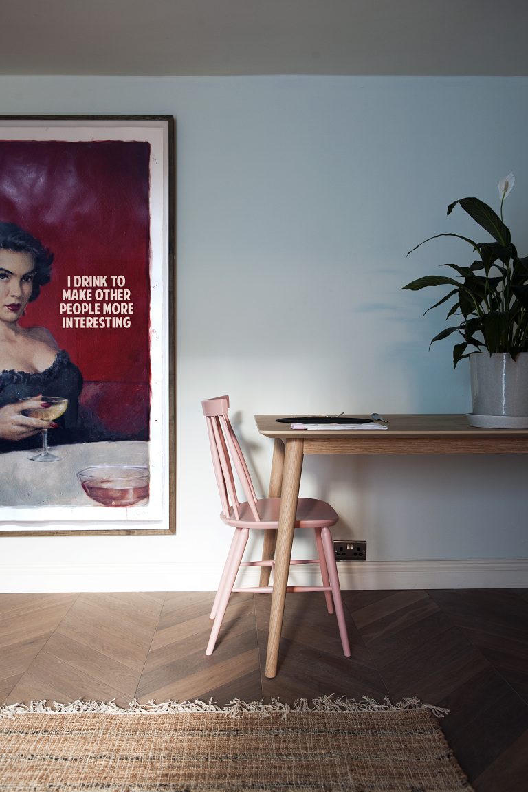 Modern interior with a pink wooden chair, minimalist desk, large houseplant, and bold wall art featuring a vintage-style woman with a drink.