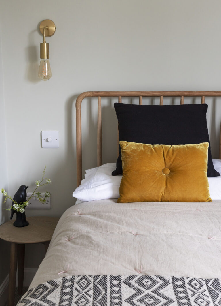 Minimalist bedroom with a wooden bed frame, mustard yellow cushion, black pillow, and a wall-mounted brass light fixture.